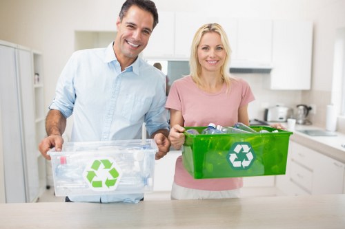 A smiling man and woman stand side by side in a bright, modern kitchen, each holding a clear recycling bin with a green recycling symbol on the front. The man, dressed in a light blue button-up shirt, holds a transparent bin filled with plastic bottles and containers. The woman, wearing a pink t-shirt, holds a solid green bin containing various recyclable waste items. The kitchen backdrop features white cabinets, a countertop with kitchen appliances, and a clean, light-colored environment. This scene depicts responsible waste segregation and recycling practices, relevant to rubbish removal services offered by House Clearance Cranford in the Cranford area, which includes surrounding towns and postal codes. The focus is on environmentally conscious waste management in a domestic setting, emphasizing proper disposal of recyclable materials across residential properties.
